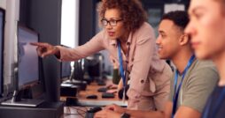 Instructor in glasses leans over student at computer, pointing at screen in classroom with other learners seated nearby.