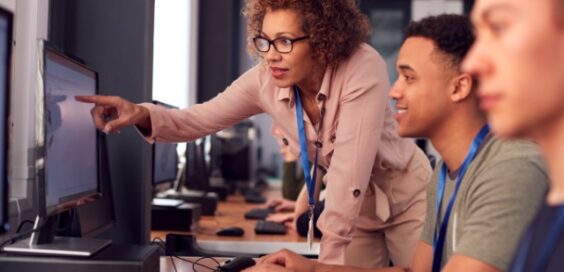 Instructor in glasses leans over student at computer, pointing at screen in classroom with other learners seated nearby.