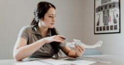 Woman seated at desk examines small airplane model, holding pen over parts while working with notebook and tools.