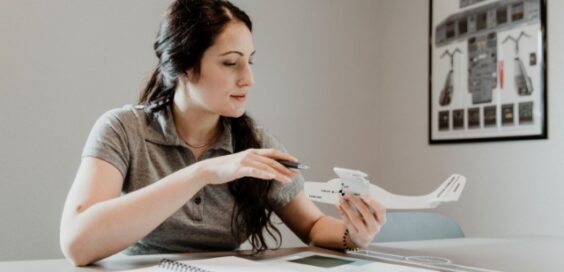 Woman seated at desk examines small airplane model, holding pen over parts while working with notebook and tools.