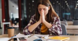 A young person prays at a table in a public space. A coffee cup, papers, a phone, and a notebook are on the table.