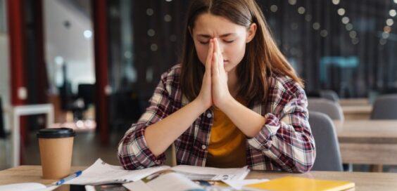A young person prays at a table in a public space. A coffee cup, papers, a phone, and a notebook are on the table.