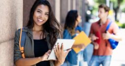 A smiling college student leaning against a building on the campus while using a small silver tablet.