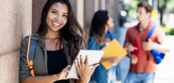 A smiling college student leaning against a building on the campus while using a small silver tablet.