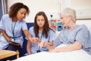 Nurse points to elderly patient’s hand while explaining care, with another nurse observing in hospital room.