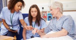 Nurse points to elderly patient’s hand while explaining care, with another nurse observing in hospital room.