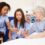 Nurse points to elderly patient’s hand while explaining care, with another nurse observing in hospital room.