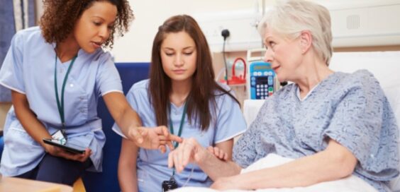 Nurse points to elderly patient’s hand while explaining care, with another nurse observing in hospital room.