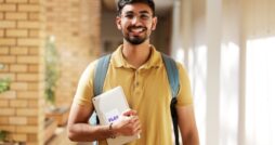 A smiling young man wearing a yellow polo shirt and a backpack is carrying an iPad against his chest.
