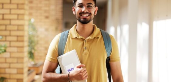 A smiling young man wearing a yellow polo shirt and a backpack is carrying an iPad against his chest.
