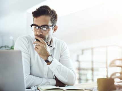A man sitting at a desk with a laptop, resting his hand on his face while looking at a screen in an office.