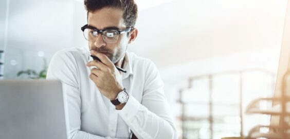 A man sitting at a desk with a laptop, resting his hand on his face while looking at a screen in an office.