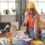 Person in a yellow beanie packs a backpack at a cluttered desk with headphones, notebooks, glasses, and a water bottle.