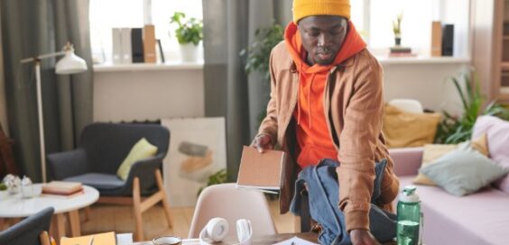 Person in a yellow beanie packs a backpack at a cluttered desk with headphones, notebooks, glasses, and a water bottle.