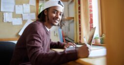 A smiling male student sits at a desk in front of a window. One hand holds a pen, and the other sits on a laptop keyboard.