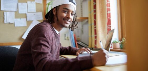 A smiling male student sits at a desk in front of a window. One hand holds a pen, and the other sits on a laptop keyboard.