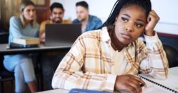 A sad college student sits at a table in a classroom, while other students sit behind her in front of a laptop.