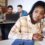 A sad college student sits at a table in a classroom, while other students sit behind her in front of a laptop.