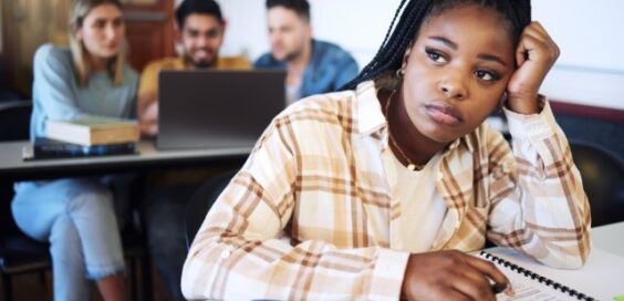 A sad college student sits at a table in a classroom, while other students sit behind her in front of a laptop.