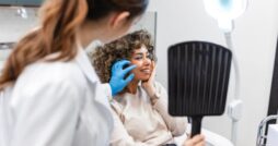 A woman holding a hand mirror while a person in a white coat and blue gloves examines her face in a clinic.