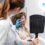 A woman holding a hand mirror while a person in a white coat and blue gloves examines her face in a clinic.