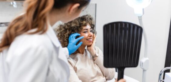 A woman holding a hand mirror while a person in a white coat and blue gloves examines her face in a clinic.