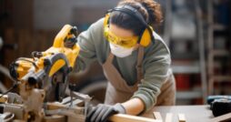 A close-up of a woman wearing a mask, safety glasses, gloves, and earmuffs while using a saw to cut wood in a workshop.