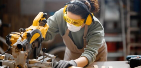 A close-up of a woman wearing a mask, safety glasses, gloves, and earmuffs while using a saw to cut wood in a workshop.
