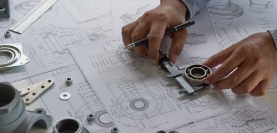 A person holds a caliper tool and a pen on top of numerous sheets of blueprints. Miscellaneous bearings are on the table.