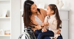 A mother in a wheelchair holding her daughter in her lap and speaking to her. They are in a white room with shelves.