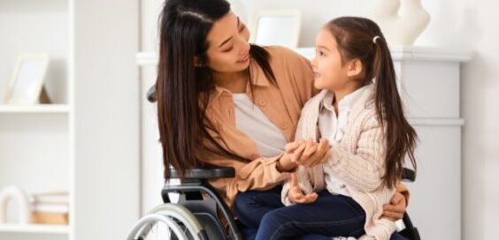 A mother in a wheelchair holding her daughter in her lap and speaking to her. They are in a white room with shelves.