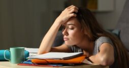 A slouching student sits with her arms on a desk and one hand on her forehead. Notebooks and a mug are on the desk.