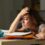 A slouching student sits with her arms on a desk and one hand on her forehead. Notebooks and a mug are on the desk.
