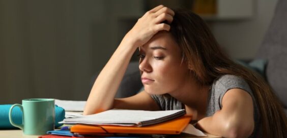 A slouching student sits with her arms on a desk and one hand on her forehead. Notebooks and a mug are on the desk.