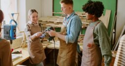 Three people wearing aprons examining a handheld power tool beside a workbench in a woodworking workshop.