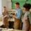 Three people wearing aprons examining a handheld power tool beside a workbench in a woodworking workshop.