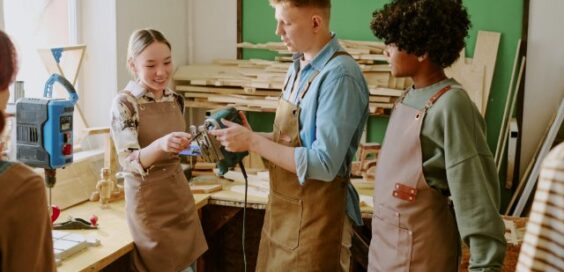Three people wearing aprons examining a handheld power tool beside a workbench in a woodworking workshop.