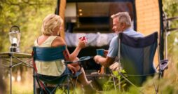 Two adults sitting in camping chairs with mugs near a small fire, an open van, grass, trees, and a lantern.