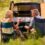 Two adults sitting in camping chairs with mugs near a small fire, an open van, grass, trees, and a lantern.