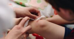 A close-up of a little kid with a knee scrape. Someone sprays something on the cut and holds a tissue near it.