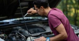 A man in a burgundy shirt using a wrench under an open car hood beside trees in a wooded area during daytime.