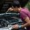 A man in a burgundy shirt using a wrench under an open car hood beside trees in a wooded area during daytime.