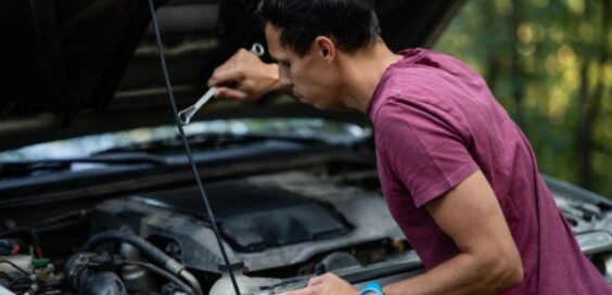 A man in a burgundy shirt using a wrench under an open car hood beside trees in a wooded area during daytime.