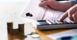 A hand holding a college financial document and pressing a calculator beside stacks of coins on a wooden table.
