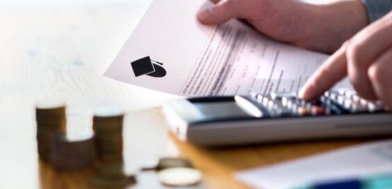 A hand holding a college financial document and pressing a calculator beside stacks of coins on a wooden table.