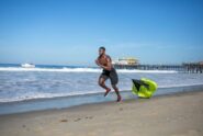 A fit man without a shirt in black shorts runs on the beach with a small parachute dragging behind him.