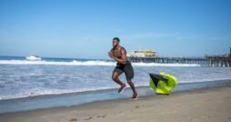 A fit man without a shirt in black shorts runs on the beach with a small parachute dragging behind him.