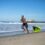 A fit man without a shirt in black shorts runs on the beach with a small parachute dragging behind him.