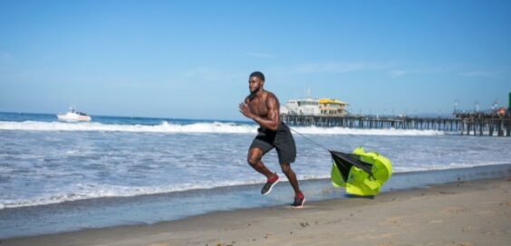 A fit man without a shirt in black shorts runs on the beach with a small parachute dragging behind him.