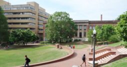 A college campus features a large brick walkway with students walking to the large brick buildings.